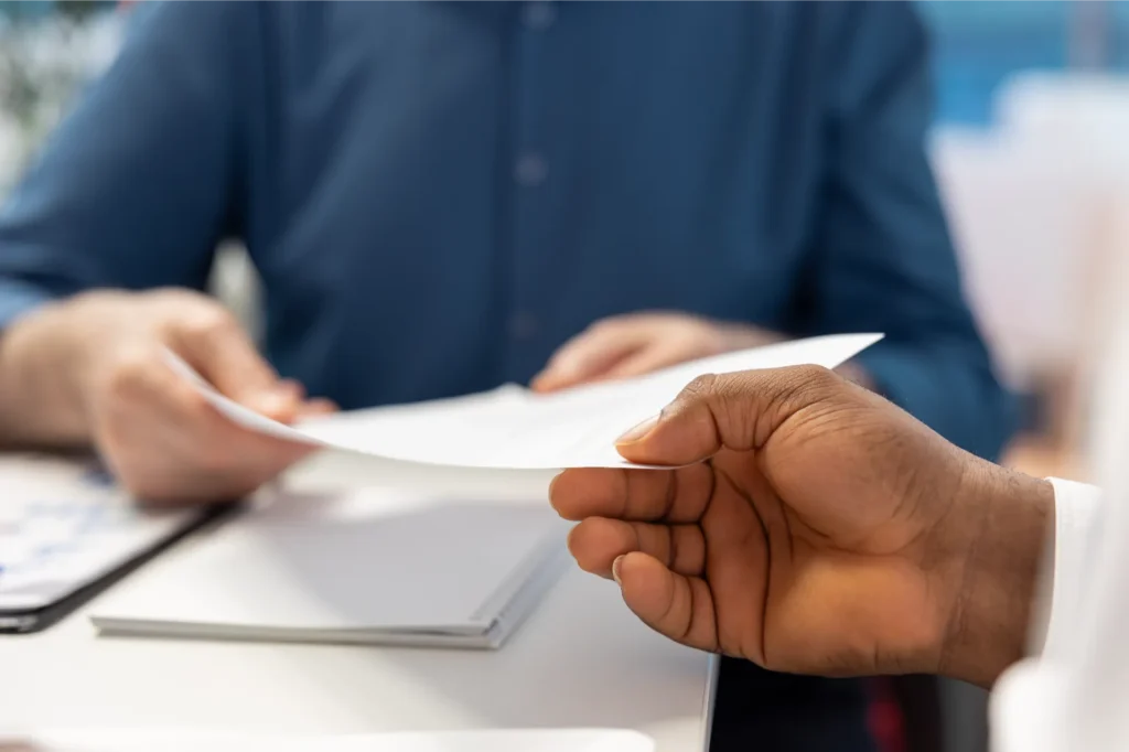 Close-up of two people exchanging a compliance report document during a business meeting.
